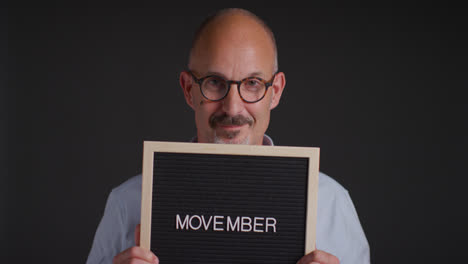 Studioporträt-Eines-Reifen-Mannes,-Der-Ein-Schild-Mit-Der-Aufschrift-„Movember“-Hochhält-Und-Das-Bewusstsein-Für-Männergesundheit-Und-Krebs-Fördert